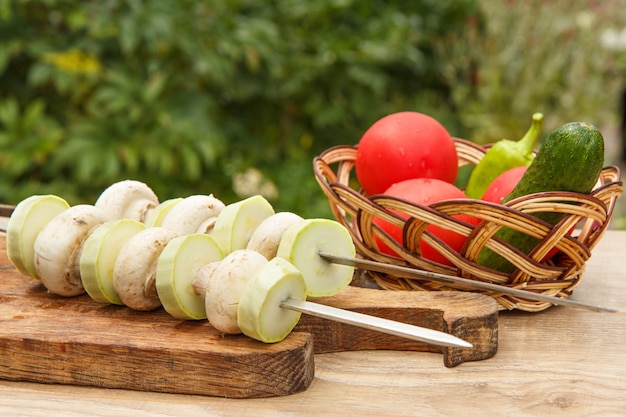 Calabacines y setas en brochetas con tomate, pepino y pimiento en una canasta de mimbre. Los calabacines y los champiñones están listos para asar con brochetas de metal.