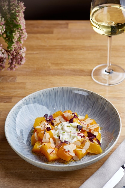 Calabaza al horno en plato de cerámica sobre mesa de madera con copa de vino blanco calabaza con stracciatella y microgreen