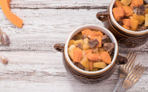 Calabaza, carne, patatas y verduras al horno en una olla de cerámica sobre una mesa de madera blanca.