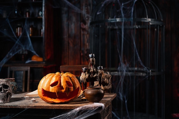 Calabaza de Halloween aterradora con cara tallada en la mesa en una habitación oscura con velas, telaraña y jaula en el fondo