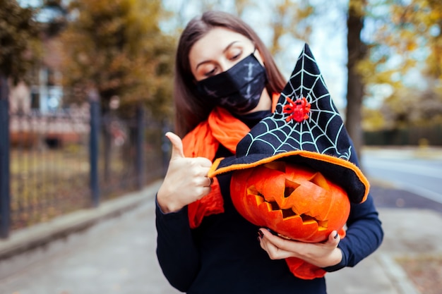 Calabaza de Halloween Jack-o-lantern. Una mujer sostiene una calabaza tallada a mano con un sombrero de bruja al aire libre con una máscara contra la pandemia del coronavirus covid-19. Celebrando seguro
