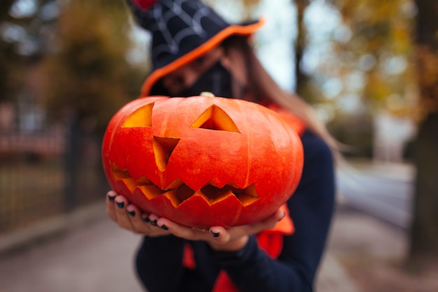 Calabaza de Halloween Jack-o-lantern. Scary girl sostiene una calabaza tallada al aire libre con una máscara contra la pandemia del coronavirus covid-19 y el sombrero de bruja. De cerca