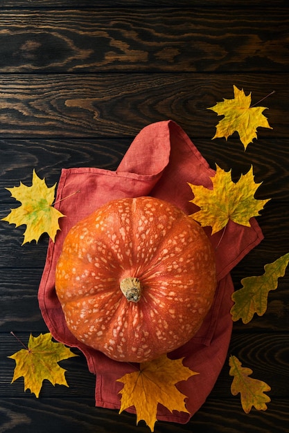 Calabaza naranja sobre una servilleta de terracota rodeada de hojas caídas sobre una superficie de madera, luz natural, vista superior, espacio para copiar