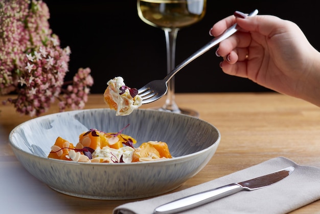 Calabaza con stracciatella y microgreen sobre mesa de madera. Mujer mantenga horquilla con calabaza y copa de vino blanco sobre fondo