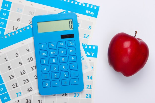 Calculadora con las hojas del calendario mensual, manzana roja sobre blanco. Cálculo económico, costeo