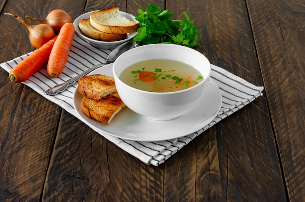 Caldo fresco en un recipiente blanco con hierbas finamente picadas con perejil y ajo sobre una mesa de madera. Sopa caliente a base de carne de pollo, zanahoria, cebolla, ajo, perejil y pimiento.