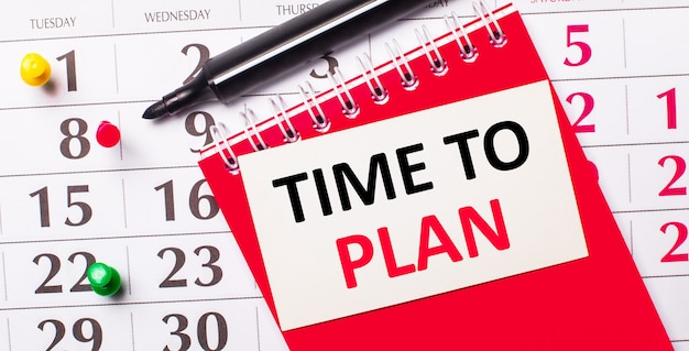 En el calendario hay una tarjeta blanca con el texto HORA DE PLANIFICAR. Cerca hay un bloc de notas rojo y un marcador. Vista desde arriba.