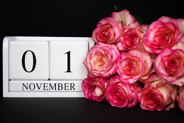 Calendario de madera del 1 de noviembre, blanco sobre fondo negro, rosas rosadas cerca.