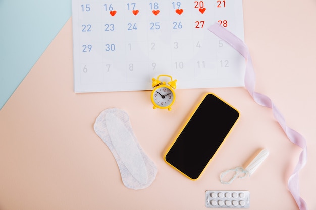 Foto calendario de menstruación y smartphone con tampón de algodón, toalla sanitaria y despertador amarillo sobre fondo rosa azul. días críticos de la mujer, protección de la higiene de la mujer