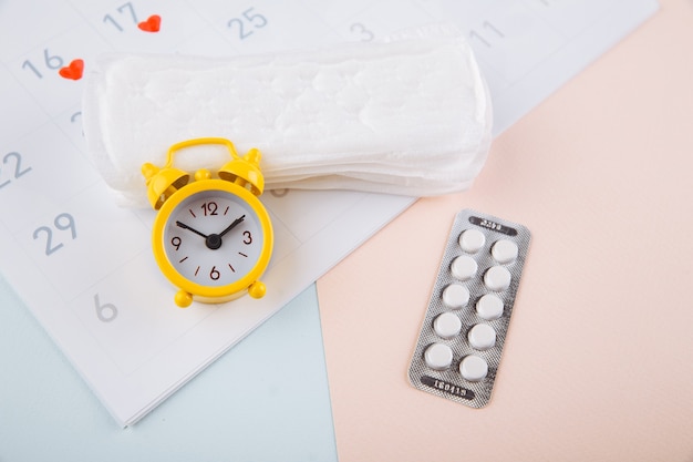 Calendario menstrual con almohadillas, reloj despertador y píldoras anticonceptivas. Concepto de período de menstruación. Analgésico para el dolor menstrual.
