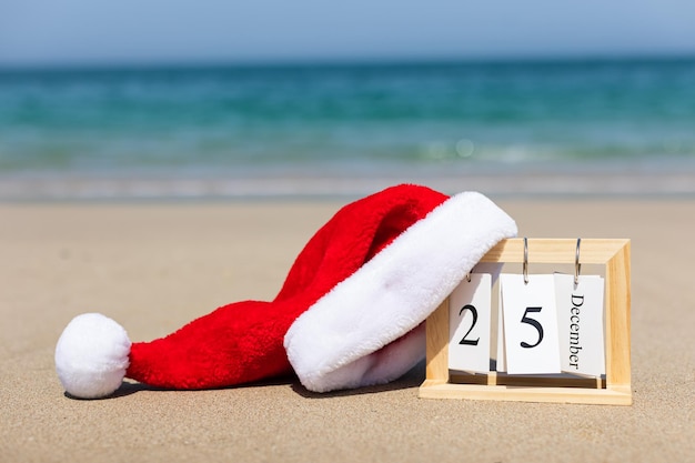 Foto calendario navideño de madera fecha 25 de diciembre en la arena con un gorro de papá noel en el verano en el hermoso mar fondo de tarjeta de navidad