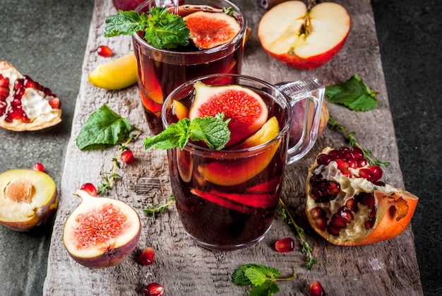 Calentamiento de otoño, invierno cócteles bebidas recetas. Sangría de fruta roja caliente con manzanas, ciruelas, higos, granada, menta, canela, tomillo, limón. En la mesa de piedra oscura, con tabla de cortar de madera,