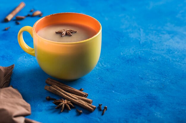 El calentamiento popular asiático bebe té de masala en una taza amarilla con cilantro, canela y tallo de rosa sobre un fondo azul clásico.