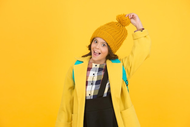 Cálido y elegante. Feliz pequeña colegiala toque elegante sombrero de pompón. Sonrisa de niño pequeño con elegante look otoñal sobre fondo amarillo. Elegante y confortable.