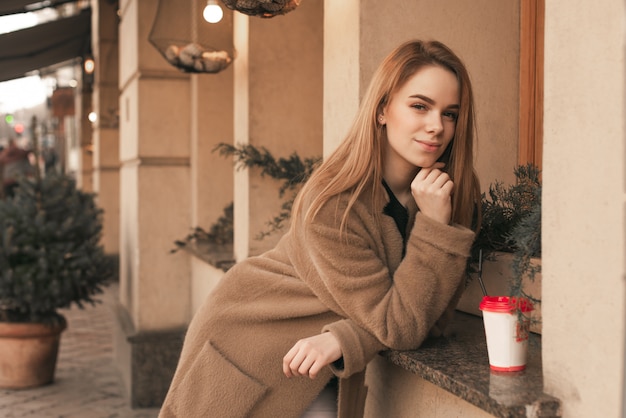 Calle retrato de una joven con un abrigo, de pie en el fondo de una pared de color beige en el alféizar de la ventana de un restaurante con una taza de café