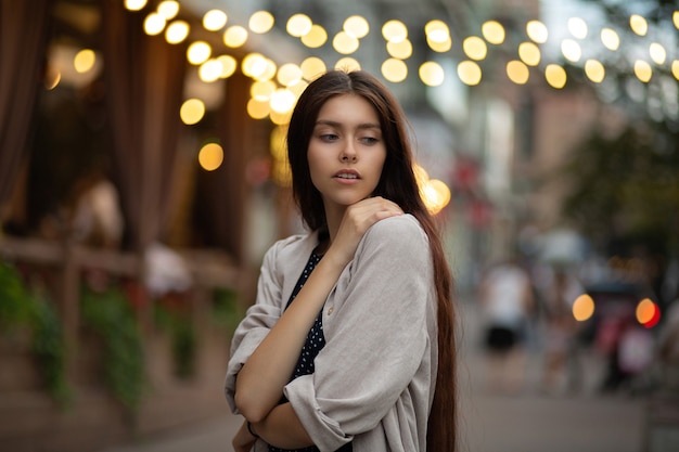 Calle retrato de mujer joven atractiva viste camisa beige posando en las luces de la ciudad por la noche