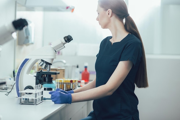Calma assistente de laboratório sentada à mesa com tubos de ensaio e um microscópio moderno à sua frente