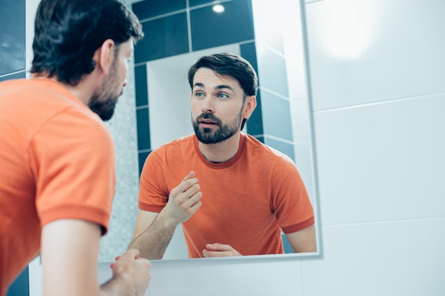 Foto calma e atraente jovem de camiseta laranja em pé no banheiro em frente a um espelho e olhando para seu reflexo