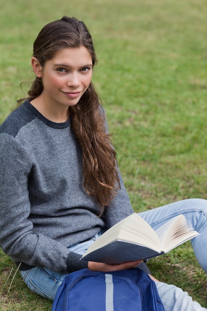 Calma estudante segurando um livro enquanto está sentado ao lado de sua mochila