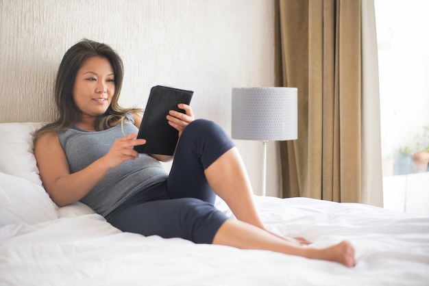 Calma hermosa mujer japonesa leyendo un libro electrónico en la cama. Chica en top gris acostado en la cama en posición acogedora con tableta