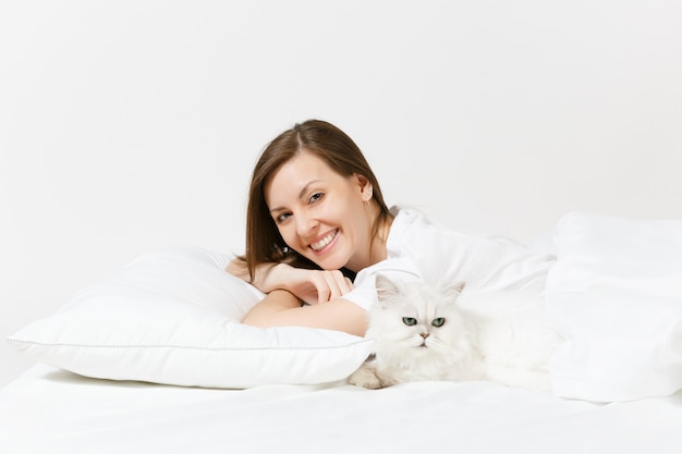 Calma jovem deitada na cama com um lindo gato persa branco de chinchila prateada, lençol, travesseiro e cobertor na parede branca