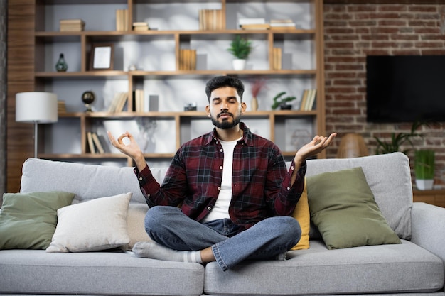 Calma de un joven barbudo haciendo una pose de loto de yoga para meditar y relajarse en el sofá durante el trabajo