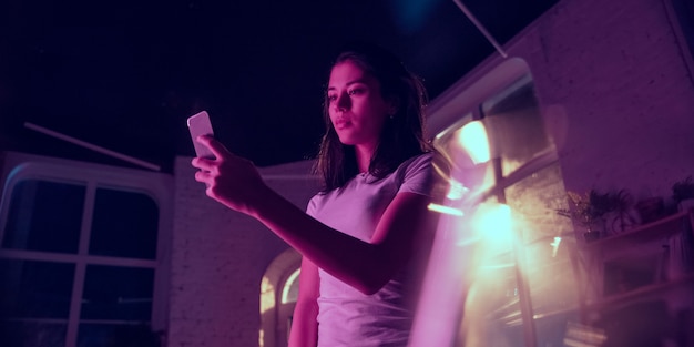 Calma. Retrato cinematográfico de uma mulher bonita e elegante no interior iluminado por néon. Tons de efeitos de cinema em azul-púrpura. Modelo feminino caucasiano usando smartphone em luzes coloridas dentro de casa. Folheto.