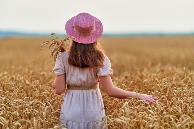 Calma, serena e livre, jovem mulher usando chapéu e vestido sozinha em um campo de trigo seco amarelo dourado e desfrutando de um belo momento de liberdade