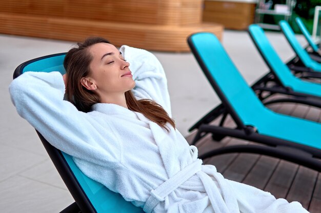 Foto calma serena mujer vestida con bata de baño con los ojos cerrados y las manos detrás de la cabeza relajándose y acostado en una tumbona en el balneario de bienestar. satisfacción, bienestar y tiempo de recreación. estilo de vida fácil