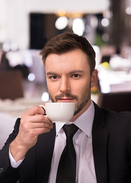 Calmado y seguro. Apuesto joven en ropa formal tomando café y mirándote mientras estás sentado en el restaurante