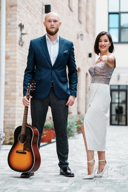 Calvo hipster barbudo con guitarra con linda mujer en vestido de noche