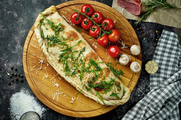 Calzone de pizza italiana apetitosa con diferentes rellenos en una composición con ingredientes sobre una tabla de madera sobre una mesa negra. Vista superior