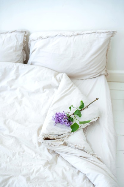 Cama con hermosas sábanas blancas arrugadas en un dormitorio blanco y flores lilas Trabajo de oficina trabajo remoto desde casa en la cama Aprendizaje a distancia educación y trabajo en línea