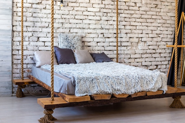 Cama de madera con almohadas y una manta de piel colgando de las cuerdas en el interior del desván de un dormitorio moderno y elegante. Áspero estilo de muebles de lujo escandinavo.