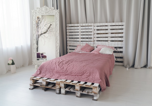 Cama Queen hecha de paletas de madera y espejo grande de madera en un dormitorio luminoso.