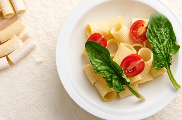 Camada plana de um macarrão de trigo integral italiano tradicional não cozido vegetariano com metades de tomate e folhas verdes de espinafre em um fundo enfarinhado com tortiglioni cru espalhado