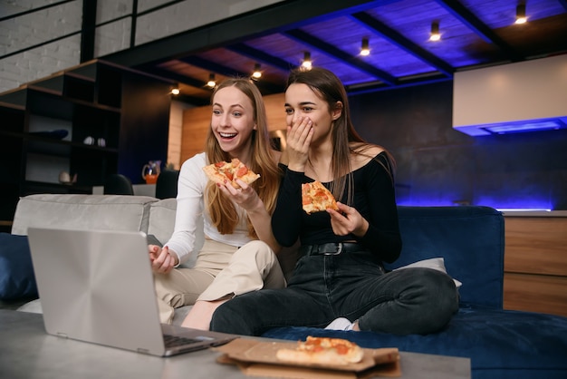 Cámara lenta de sorprendidas y atractivas amigas elegantes de 25 años que se sientan en el cómodo sofá y disfrutan de una deliciosa pizza mientras navegan imágenes divertidas en la computadora