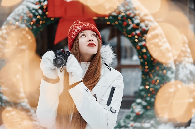 Con la cámara en las manos Mujer joven feliz de pie al aire libre y celebrando las fiestas navideñas