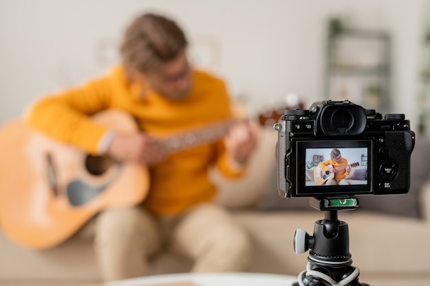 Cámara de video con músico casual joven contemporáneo tocando la guitarra en el sofá mientras graba su lección en casa para la audiencia en línea
