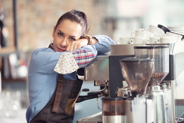 Camarera aburrida se inclina contra la máquina de café sin trabajo que hacer, sosteniendo la mascarilla en la mano.