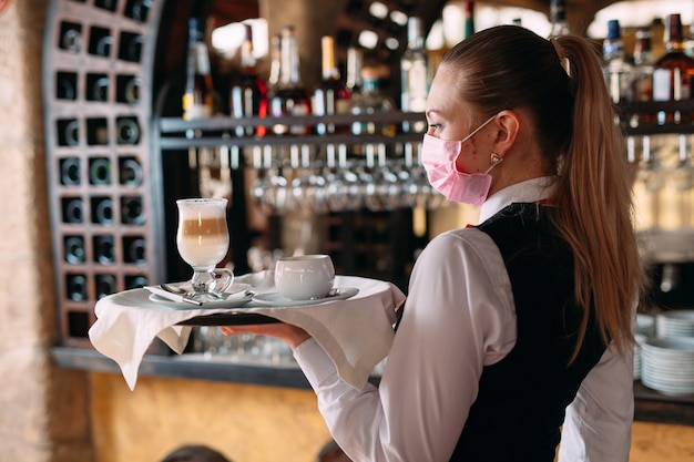 Una camarera de aspecto europeo con una máscara médica sirve café con leche.