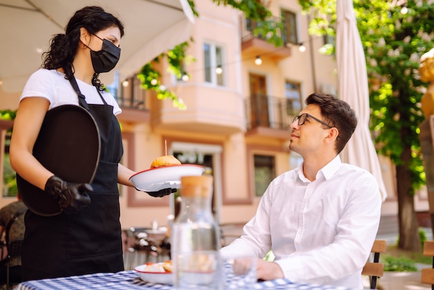 Camarera con máscara protectora que sirve comida deliciosa para el hombre en un acogedor restaurante.