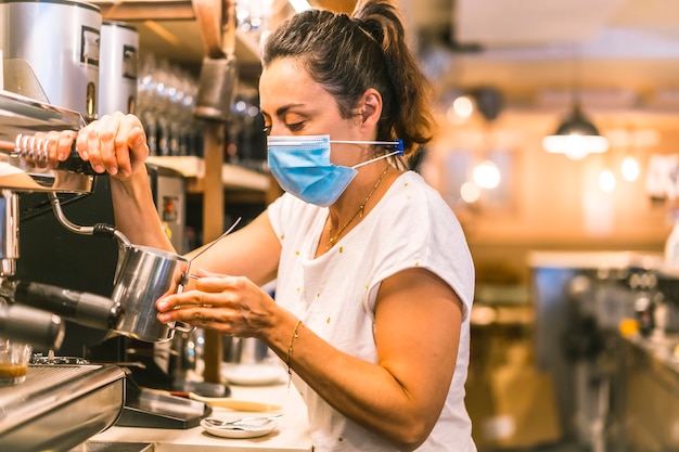 Una camarera con una mascarilla en un bar. Preparando un café con leche con la cafetera