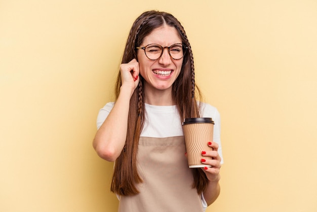 Camarera de restaurante sosteniendo un café para llevar aislado de fondo amarillo cubriendo las orejas con las manos