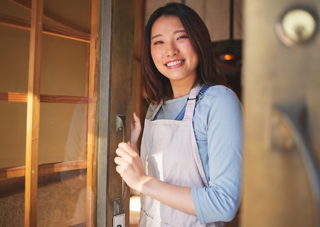 Camarera retrato y mujer asiática en la puerta de un restaurante, cafetería o tienda minorista. Sonrisa facial o barista confiado, empleado feliz o empresario de pequeñas empresas, bienvenido al inicio de una cafetería en Japón.
