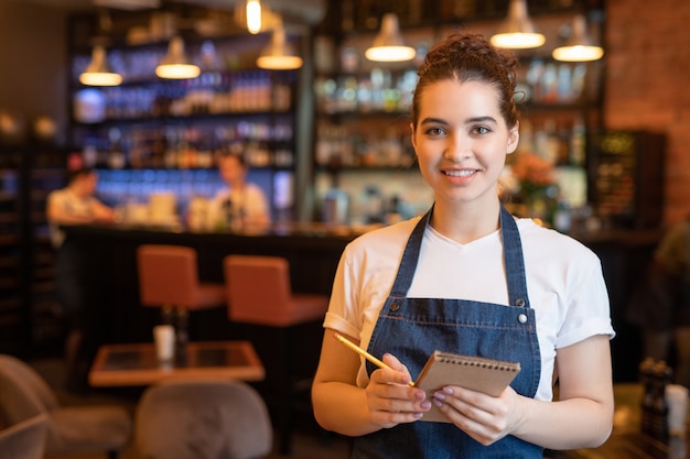 Camarera sonriente joven en ropa de trabajo de pie delante de la cámara en un restaurante de lujo y va a escribir orden del cliente