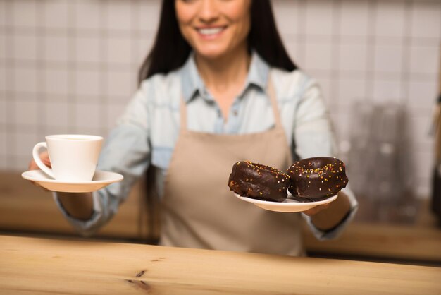 Camarera sonriente sosteniendo donuts de chocolate y una taza de café en primer plano