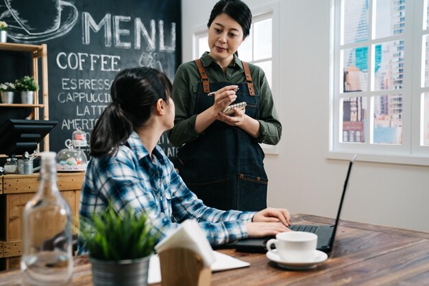 Una camarera sonriente usa delantal tomando orden en el café. una joven cliente sentada en una mesa de madera hablando con el personal en una cafetería mientras trabaja en una computadora portátil junto a la ventana en un lugar acogedor de la ciudad moderna