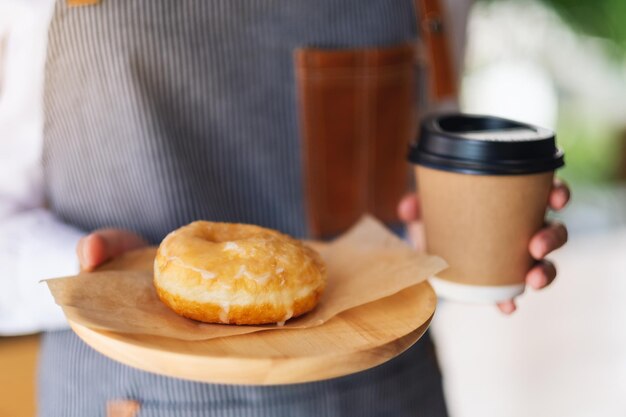 Una camarera sosteniendo y sirviendo un trozo de rosquilla casera en bandeja de madera y una taza de café de papel