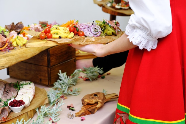 La camarera con un vestido popular rojo ruso pone una bandeja de madera con bocadillos fríos en la mesa del buffet